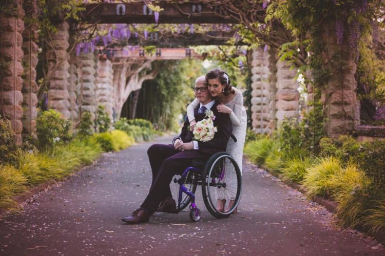 groom in wheelchair and bride hugging him from behind, under floral arrchways