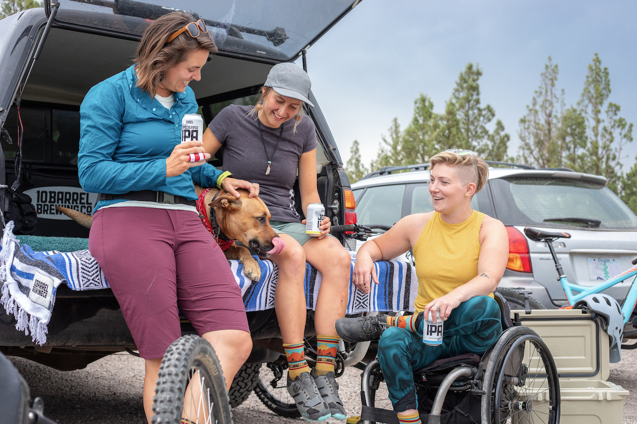 Two women sitting on the back of a truck petting a dog and drinking beer with a woman using a wheelchair next to the car