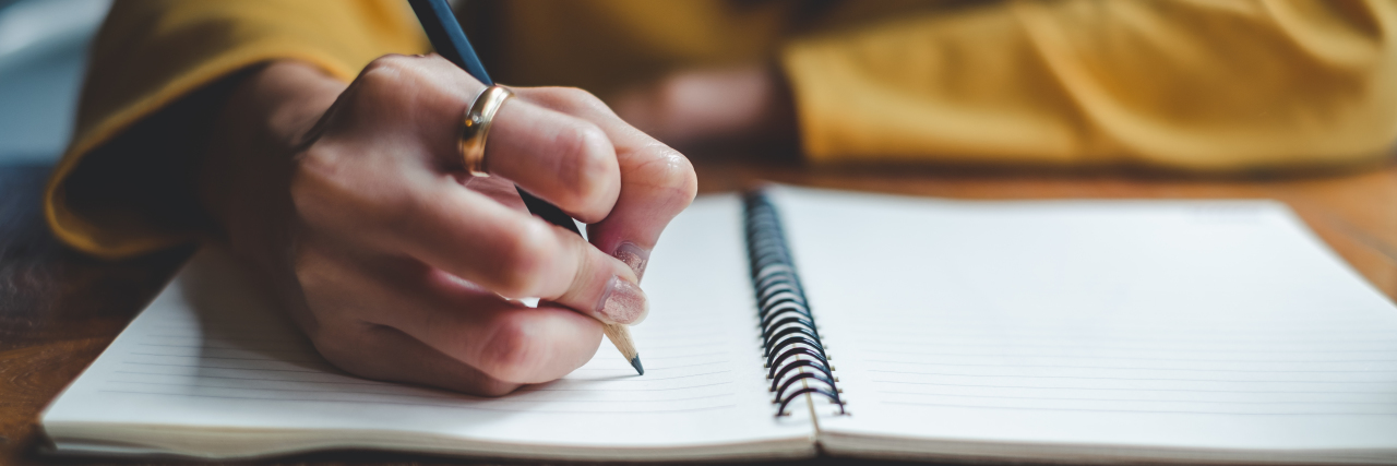 Zoomed in picture of a woman with a yellow long sleeve shirt adn a ring on her pointer finger writing in a notebook with a pencil