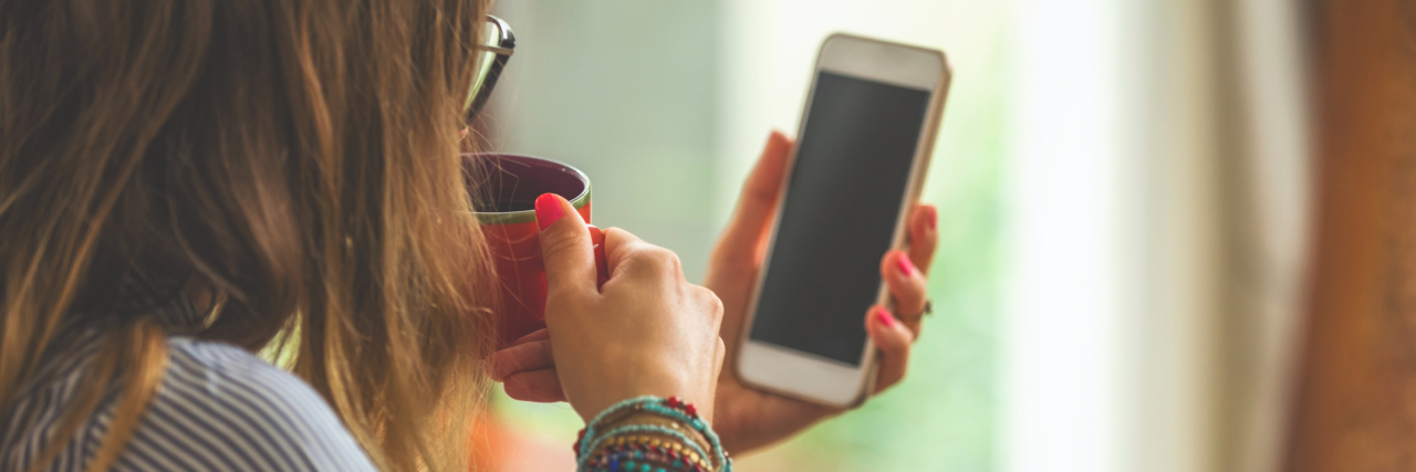 Apps Offer Therapy Resources for People Who Need Them Woman using cellphone and laptop inside house and drinking coffee / tea.