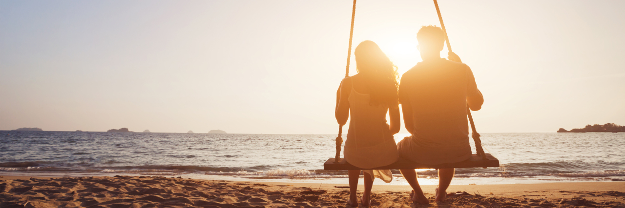 Should You Disclose Your Chronic Illness When Dating? Couple sitting together on rope swing at sunset on beach.