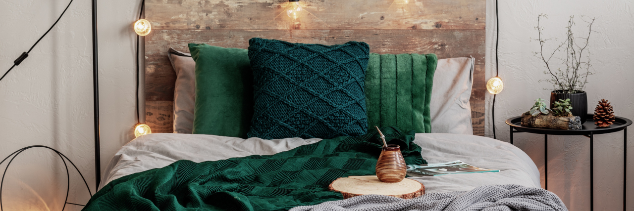Photo of bedroom. Green pillows, a green and grey blanket draped over bed. Wooden headboard with yellow string lights. Cup of tea sitting on bed