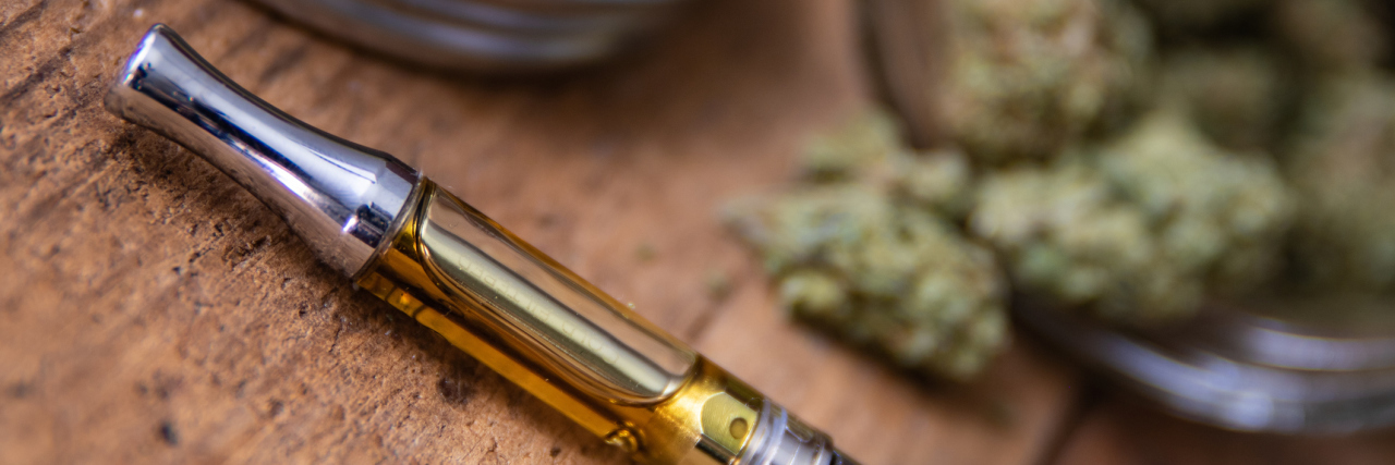 What to Know About Vape-Related Illness and CBD Products THC oil concentrate filled vape pen on natural wood with an open glass container full of marijuana buds