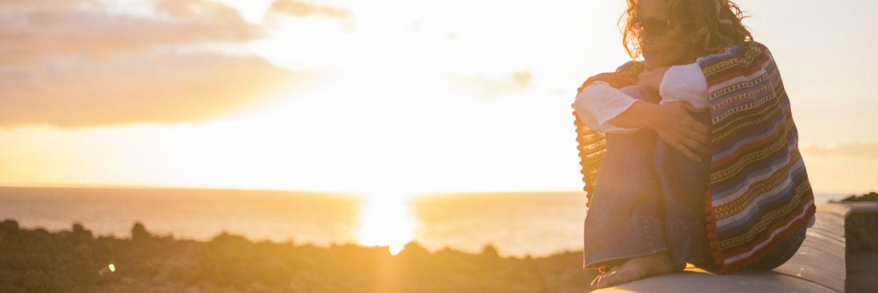 The Heaviness of My Grief After Losing My Son to Suicide Woman with curly hair, a white shirt, jeans, and hippy style blanket sitting on top of a wall with the sunset in the back