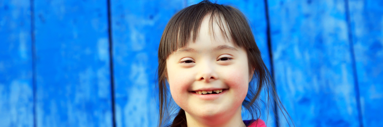 Down Syndrome Is Not Sad or Tragic Girl with Down syndrome smiling. Blue wall in background.