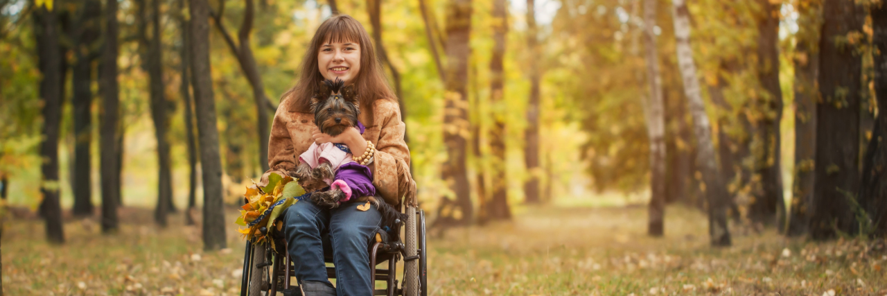 Woman in a wheelchair holding a small dog in autumn landscape.