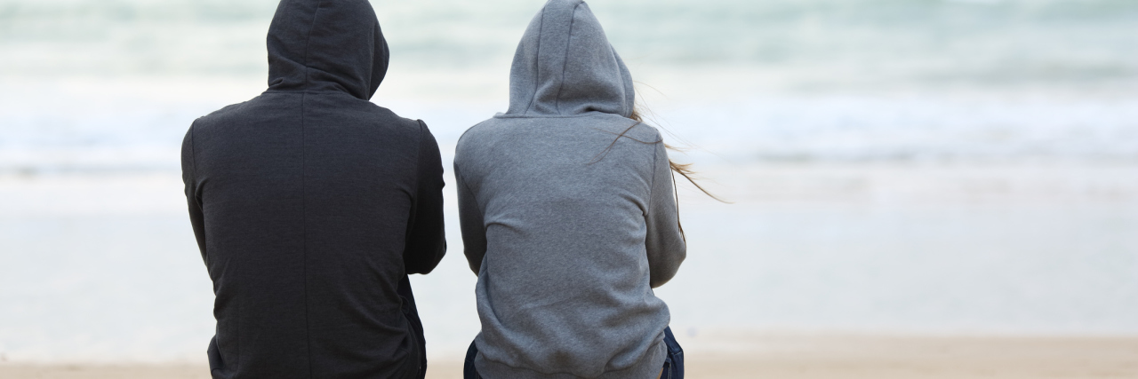 View of the back of teenagers, left in black hoodie, right in grey hoodie, sitting on a beach looking into the ocean