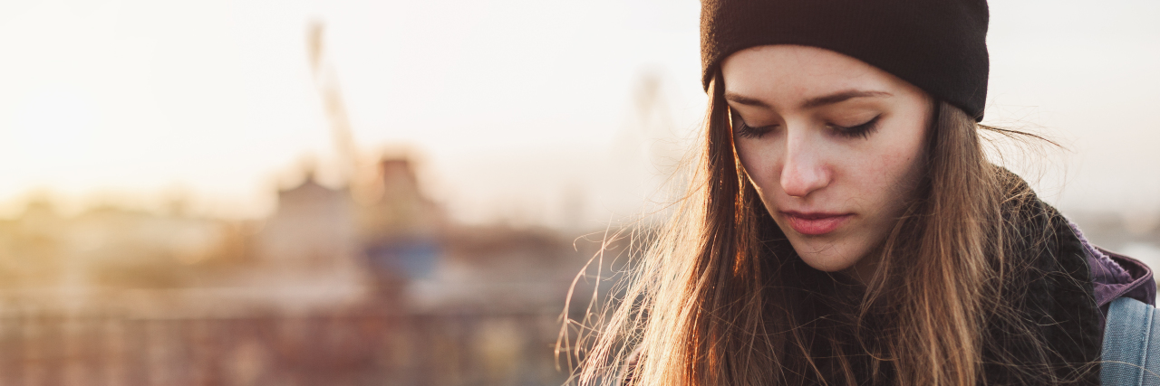 Young woman with long hair and a beanie looking down with a sunset background