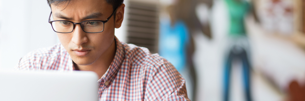 Portrait of serious young Asian student wearing glasses sitting at table and using laptop in cafe