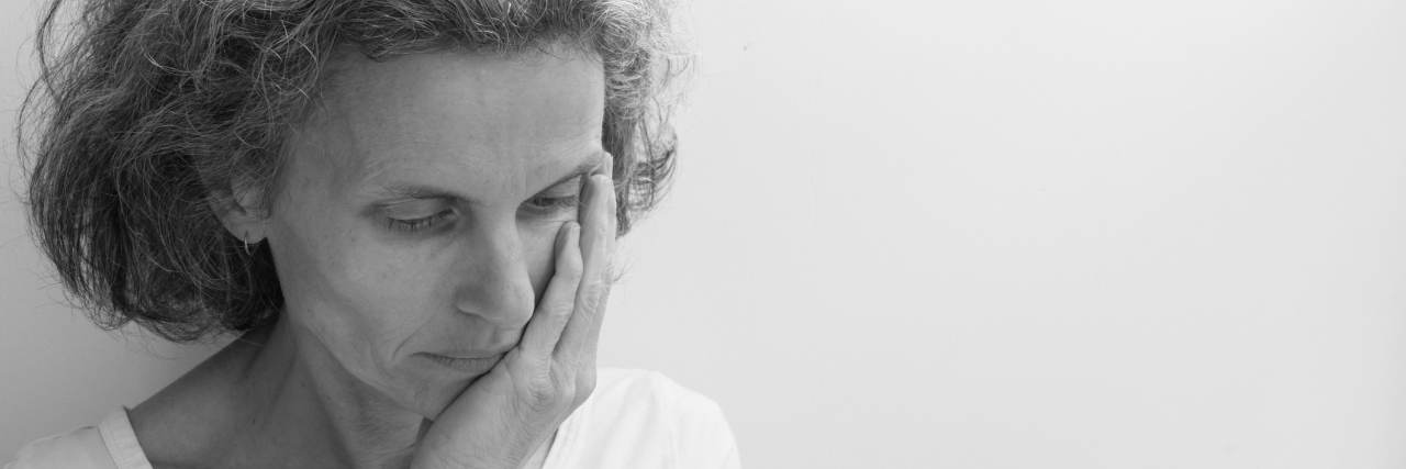 Middle aged woman in white top wiht hand on face against neutral background with copy space to right (black and white, selective focus)