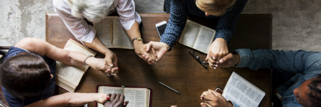 Birds eye view picture of 6 people at a table together holding hands with bibles, praying.