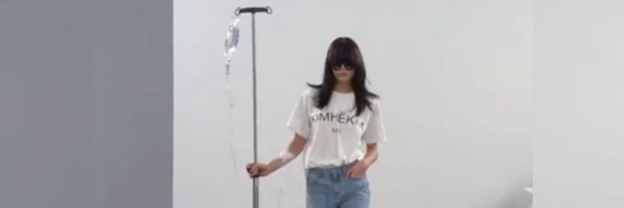 Kimhēkim model with long dark hair and sunglasses wheeling a fake IV pole