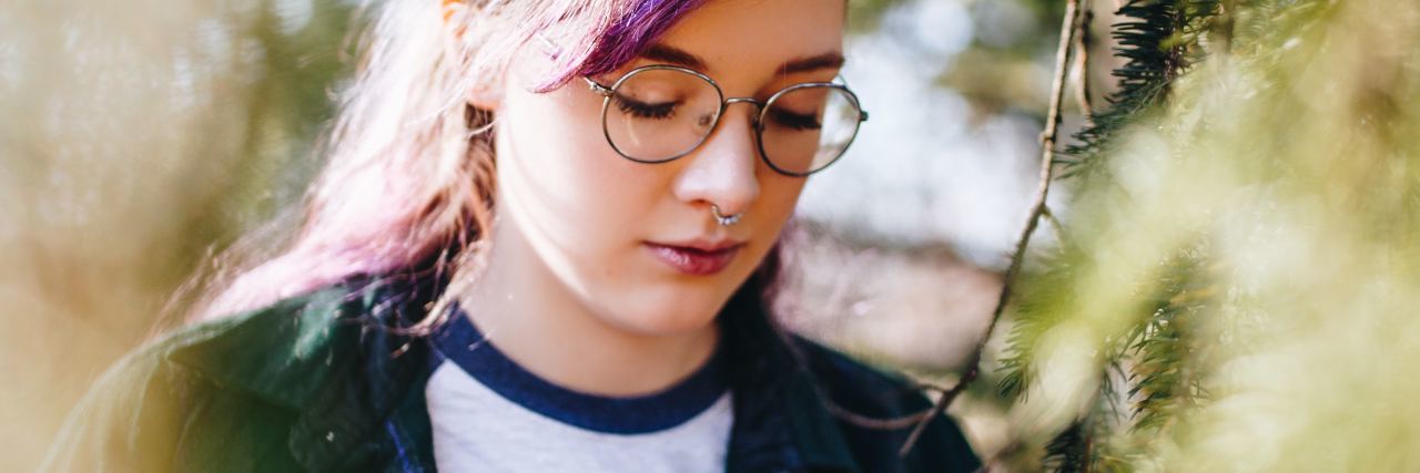 photo of young woman wearing glasses standing in trees looking down, sad