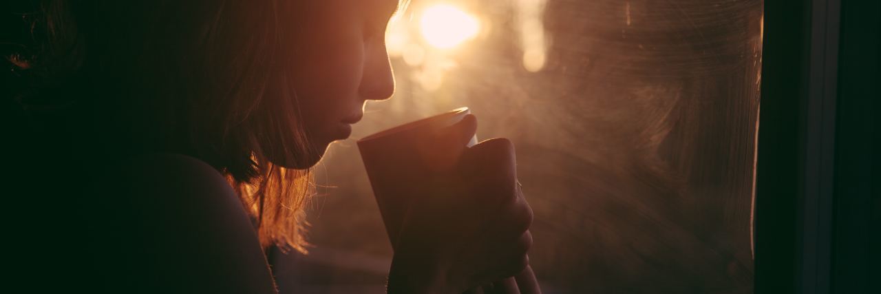 Having Mixed Feelings in Self-Harm Recovery photo of woman drinking coffee by window, silhouetted by sunset, looking down at her mug