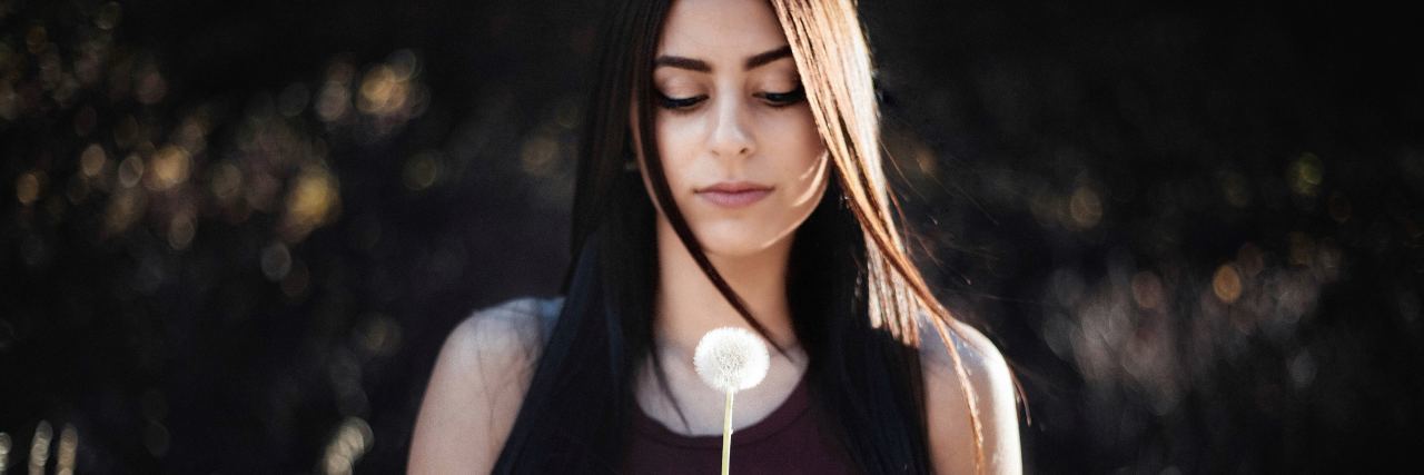 photo of woman with dark hair holding dandelion and looking down ast it
