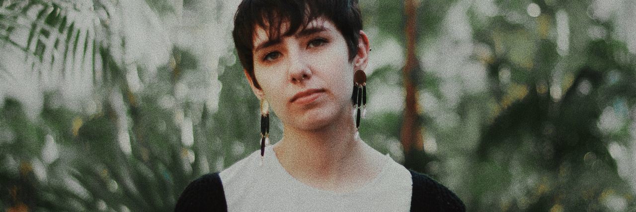 A woman with short hair and long earrings standing in front of trees