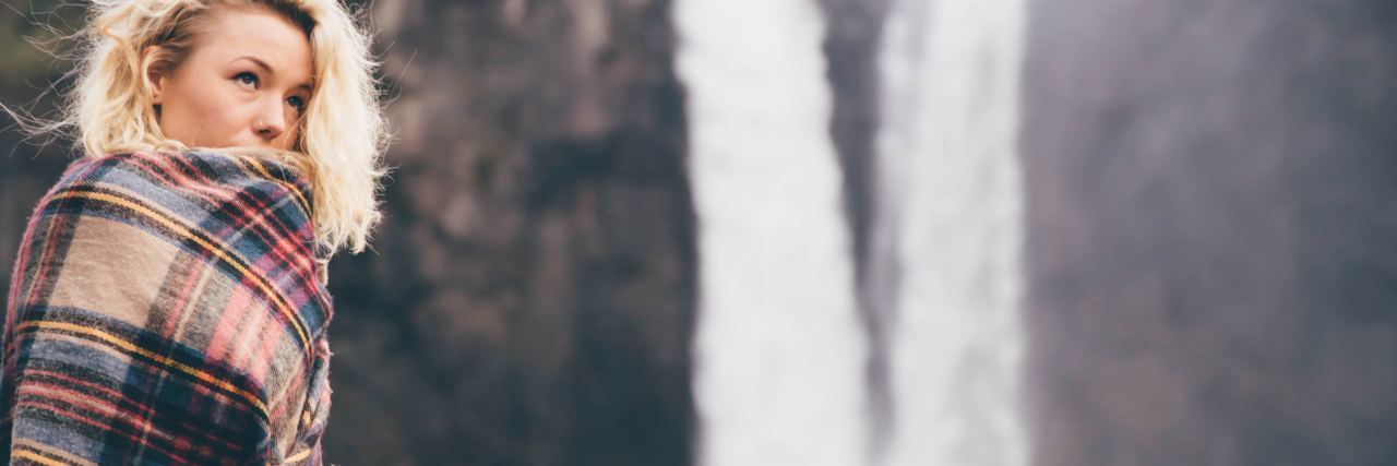 photo of woman wrapped in blanket standing beside tall waterfall