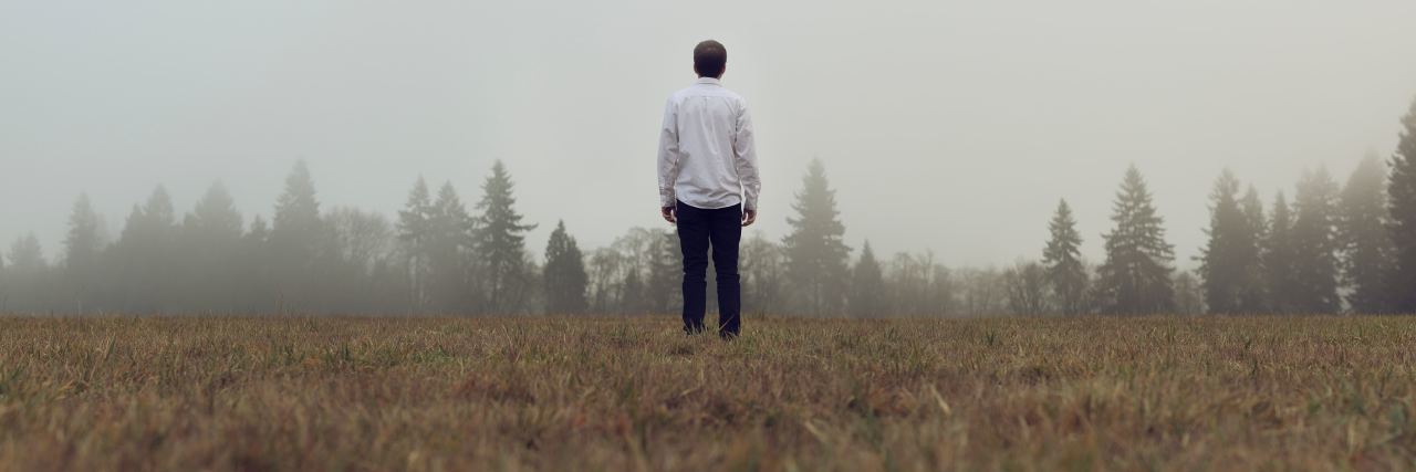 photo of man standing in misty field looking at trees in distance with back to camera