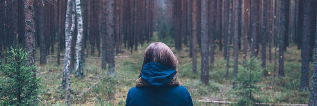 photo of woman standing with back to camera, staring into thick woods with tall trees