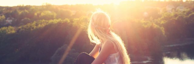 Back of a blonde girl sitting on a rock with a white top and black skirt looking out into the a forest with the sun shining down