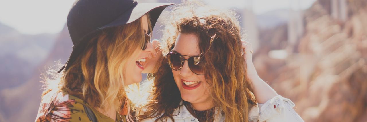 Two girls, one in a black hat, both with sunglasses laughing with each other
