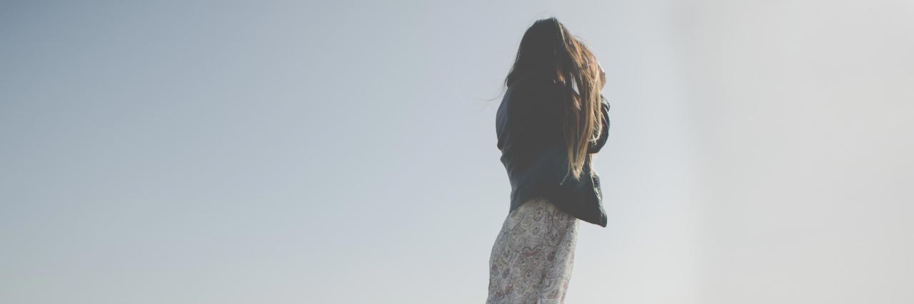 I'm Glad I Didn't Die by Suicide photo of woman standing by shore wearing dress and jacket, looking out at water