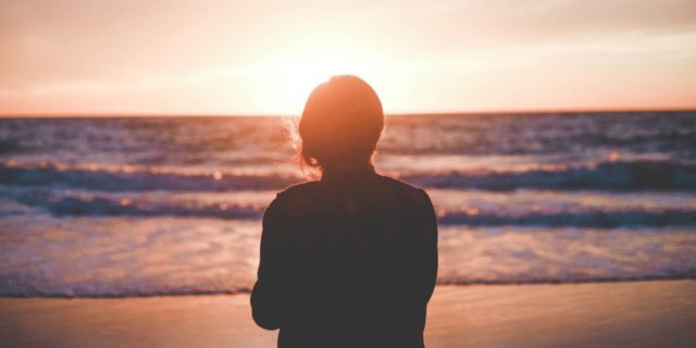 photo of woman standing on beach looking out at waves at sunrise