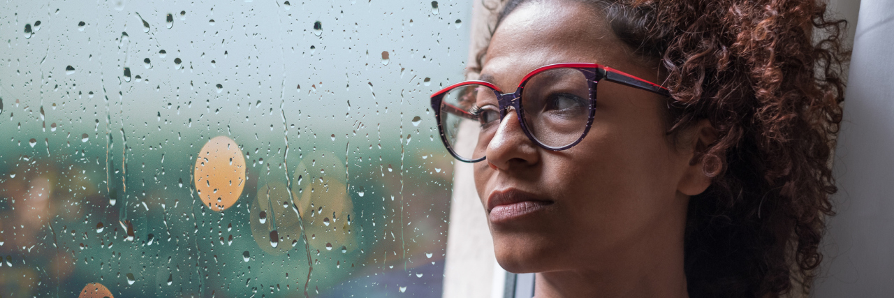 Being in a Constant State of Pain With Fibromyalgia A woman sits alone looking outside a window soaked with rain.