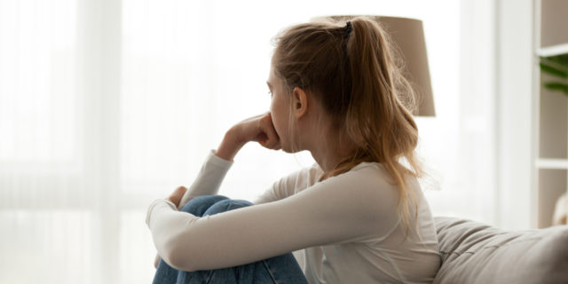side view of a woman in jeans a long sleeve white shirt sitting on a couch looking out the window