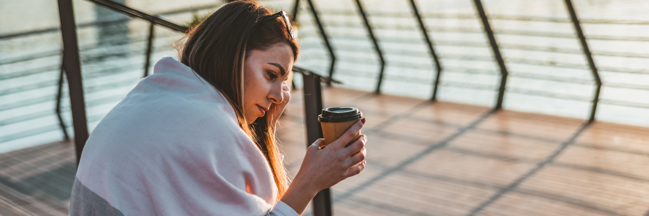 How I Found Treatment and a Good Therapist for My OCD woman sitting by the water with a white sweater and green jeans holding a cup of coffee looking down, sad