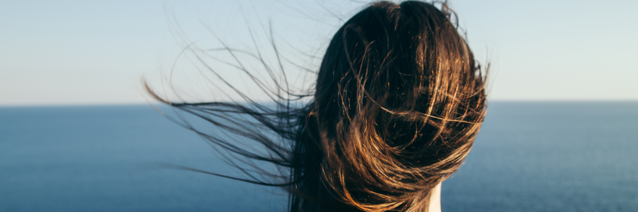 back of a woman with straight brown hair blowing in the wind as she looks out into the ocean