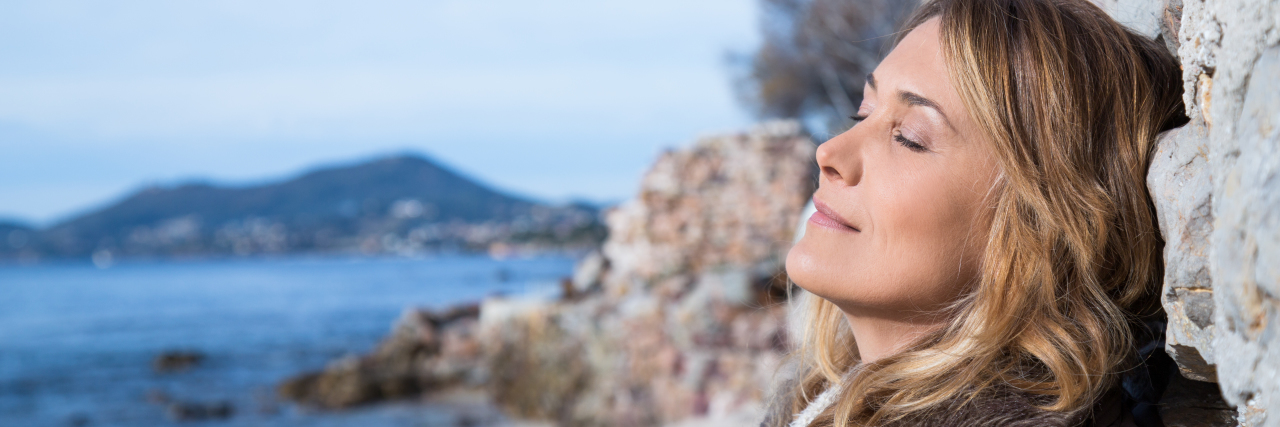 Blonde woman with her eyes closed leaning against a wall at the beach with the ocean and mountains in the background