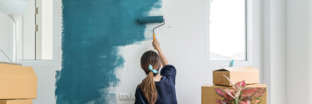 photo of young woman painting wall in home with blue paint, surrounded by boxes