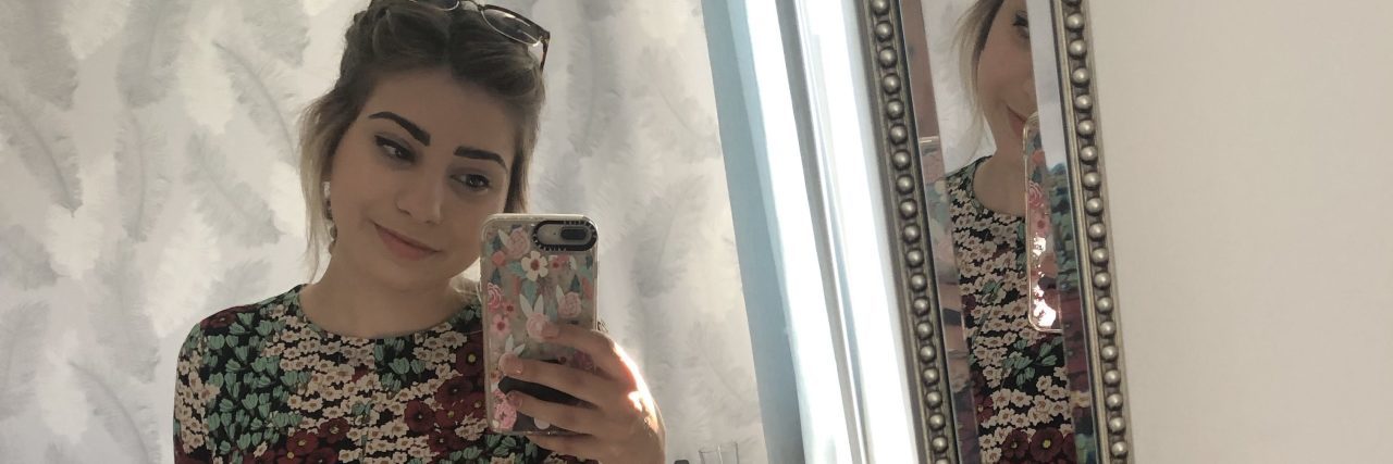 Hating Your Body With a Chronic Illness A woman takes a selfie, smiling in the mirror.