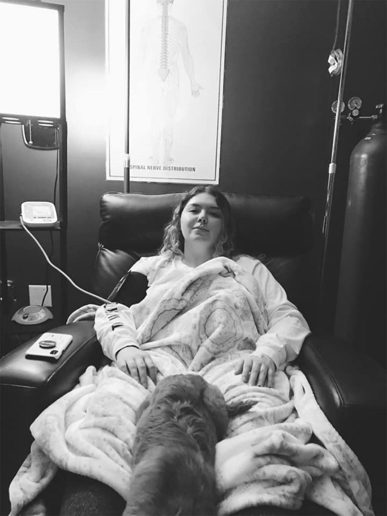 black and white photo of young woman in bed on hospital IV drip looking at camera. She also has a dog resting on her legs