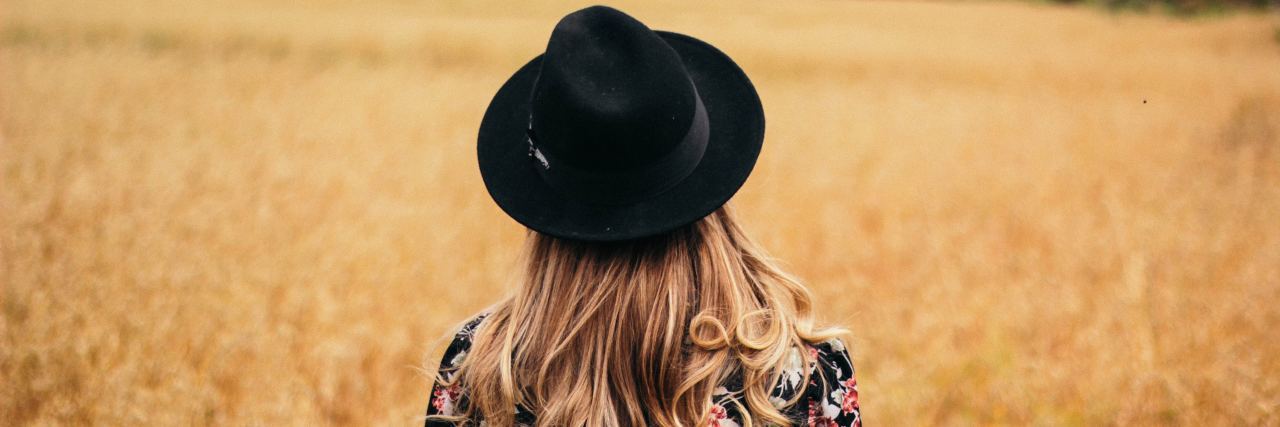 I'm Learning How to Forgive and What It Looks Like With PTSD back of woman with a black flowered shirt and black hat on with blonde hair standing in the middle of a field