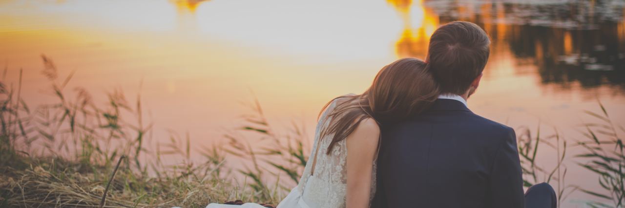 Supporting My Husband With PTSD After He Supported Me Picture of the back of a woman in a white dress with her head on the shoulder of her partner in a suit sitting on the grass looking out into a lake during sunset
