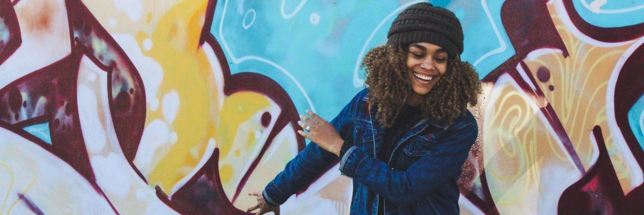 How I'm Navigating My Recovery From Depression A woman in a jean jacket and black hat dancing in front of a painted wall smiling