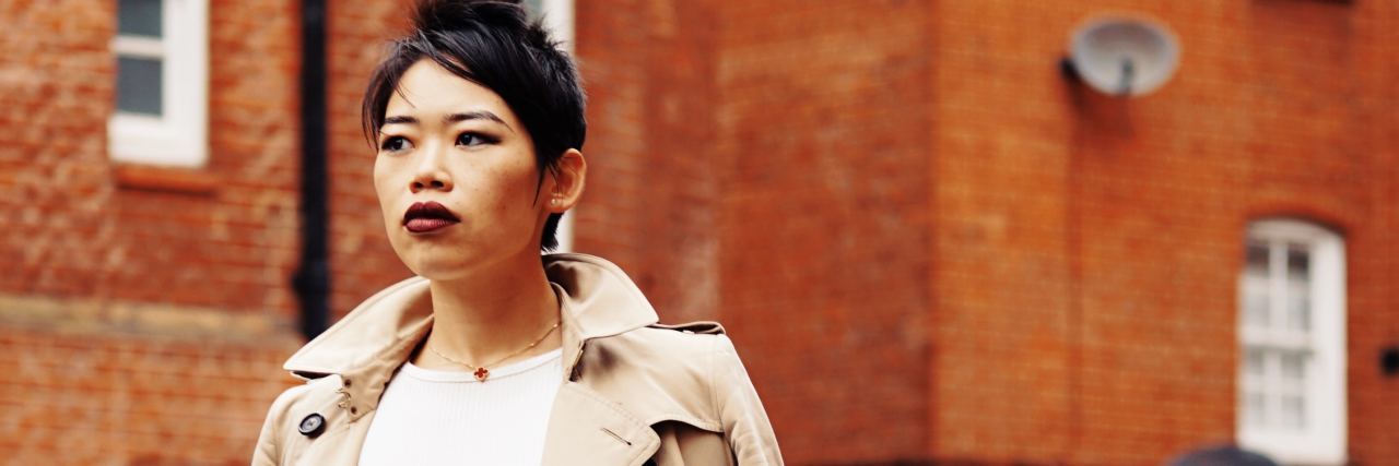 Needing to Cry With Bipolar Disorder Doesn't Make Me Weak Serious woman with red lipstick in a tan coat and black skirt facing camera in front of an orange building
