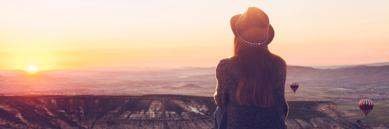 a girl with a hat sitting watching the sunrise above mountains