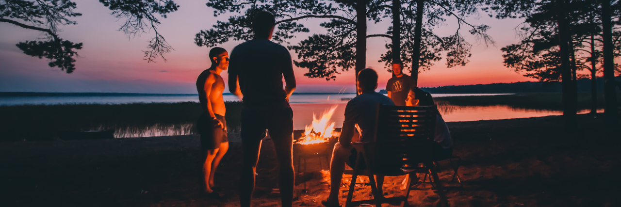group of people sitting and standing around a campfire with a lake in the background at a sunset