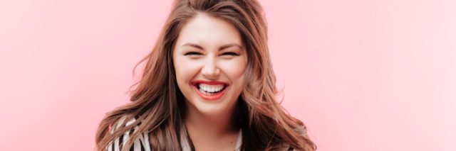 happy woman in a black and white striped blouse squinting and laughing with brown hair to her shoulders a pink background