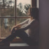A woman sits alone, sadly looking out of a window.