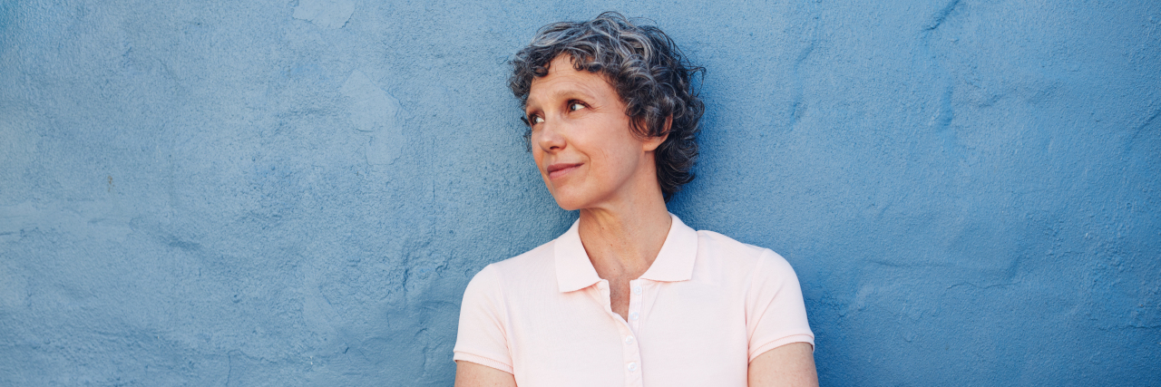 Reflecting on My Mental Illness Journey for My High School Reunion older woman against a blue backdrop with short gray hair looking up with her arms crossed