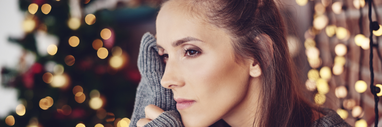 5 Tips for Navigating the Holidays If Being Around Family Is Hard woman in oversized gray sweater with brown hair looking to the side with a Christmas tree in the background