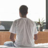 Young male is sitting on his bed with relaxation at home and looking at window. Focus on his back. Copy space in right side