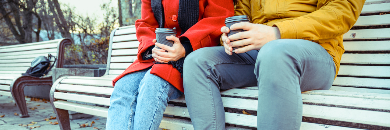 photo of 2 people sitting on a bench in fall clothes with coffee in their hands