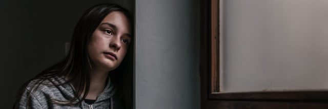 A young woman stands looking sad with her head resting against the wall.
