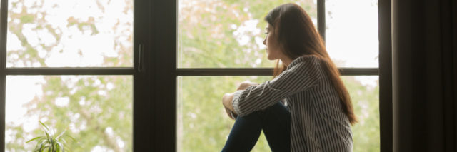 A woman sits clutching her knees looking solemnly out the window.