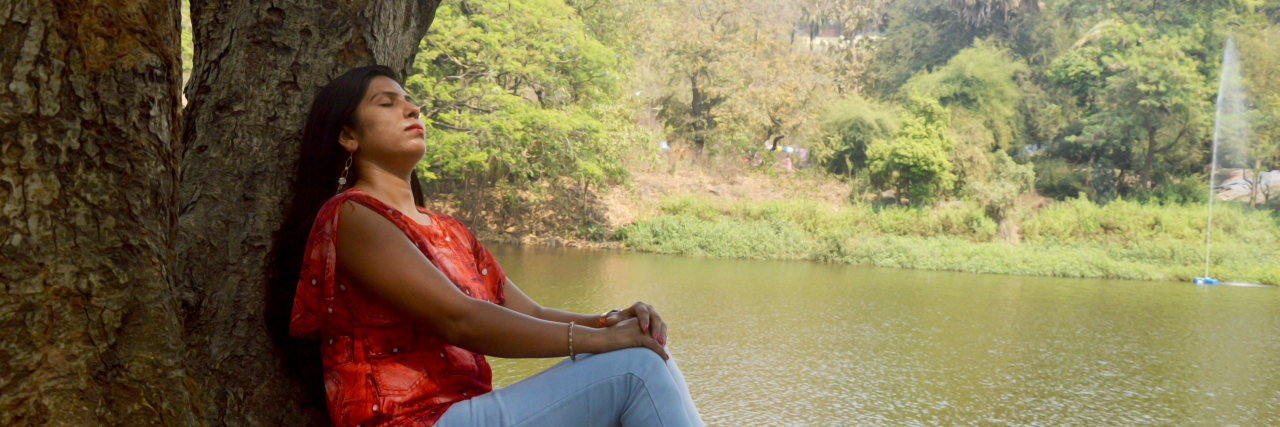 My Experience With Depression as a Teenager in India Indian woman sitting against a tree in a red shirt and blue jeans looking up with her eyes closed against the backdrop of a lake